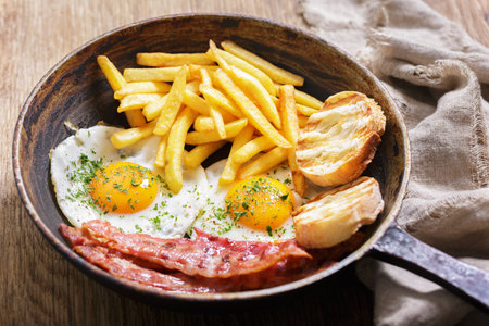 Breakfast with pan of fried eggs, bacon, toasts and french fries on wooden tableの写真素材