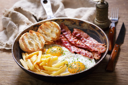 Breakfast with pan of fried eggs, bacon, toasts and french fries on wooden tableの写真素材