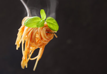 Pasta with tomato sauce and green basil on a fork on dark backgroundの写真素材