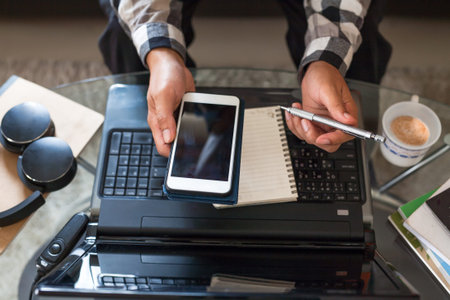 Asian man holding a smartphone and pencil to prepare meeting by using a video conference on laptop...