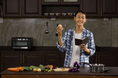 Young asian man teaching a cooking online, talking to a camera while holding some material at home kitchenの写真素材