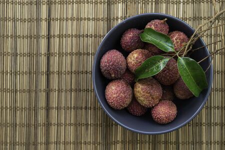 Lychee in black bowl over brown bamboo mat. Top view with copy space. Red tropical fruit with white juicy flesh.の写真素材