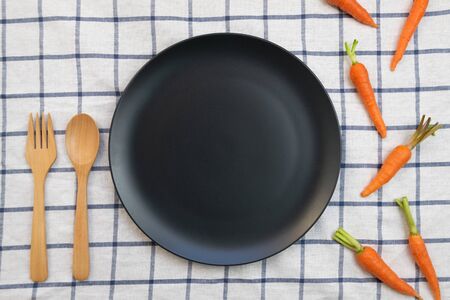Flat lay, top view of empty black dish with wooden spoon and fork and baby carrots over white table cloth with blue grid pattern. Healthy living.の写真素材