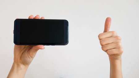 Caucasian female holds a black mobile phone with right hand and gives thumbs up with left hand on a...
