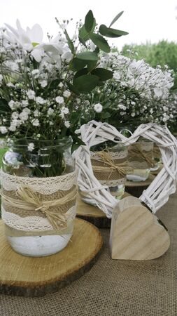 Retro decoration of the table with white daisy bouquets in glass jars, wooden heart box and wattle white heart in the gardenの写真素材