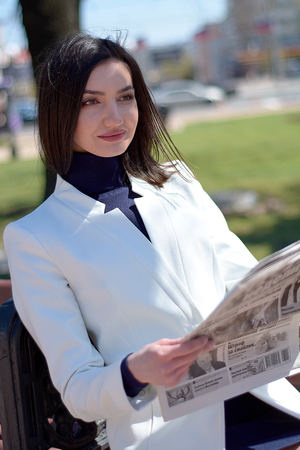 Elegant young woman dressed in a short white coat posing on a city street with a newspaper in her hands. Brunette woman. Modern urban woman portrait. Fashion business style clothes.の写真素材