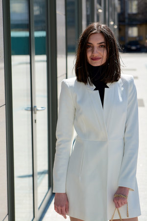 Elegant young woman dressed in a short white coat posing on a city street. Beautiful brunette woman....