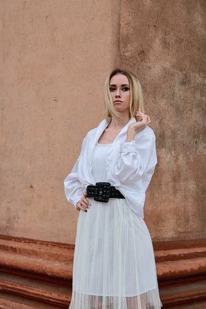 Fashion look's woman. Young woman modern portrait. Young woman dressed in white skirt and shirt posing near the old looking soviet union's architectural building with large pillars and bas-reliefs.の写真素材