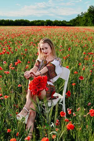 Long-haired blonde young woman in a white short dress on a field of green wheat and wild poppies. Slim sexy girl. A concept of freedom and freshness. Summer vacation female symbol. Summertime flowers.の写真素材