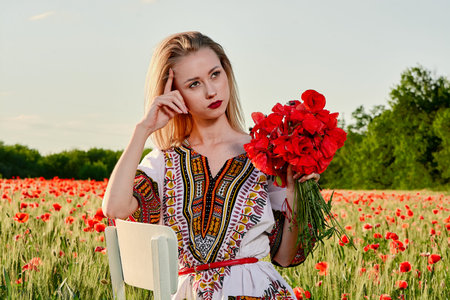 Long-haired blonde young woman in a white short dress on a field of green wheat and wild poppies....
