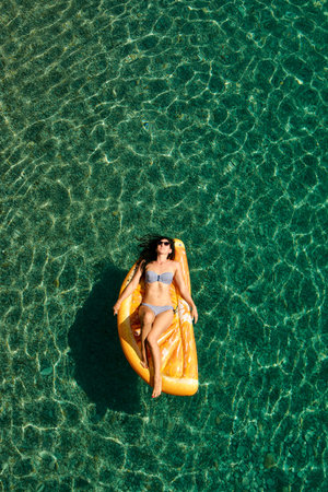 Slim young woman in bikini and sunglasses on the air mattress floats in the open sea. Enjoying...