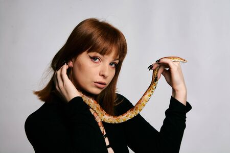 Beautiful woman in black bodywear and snake. Ginger model girl with fashion perfect make up. High end retouch. White background. Young woman with perfect fresh skin. Woman with snake around her.の写真素材