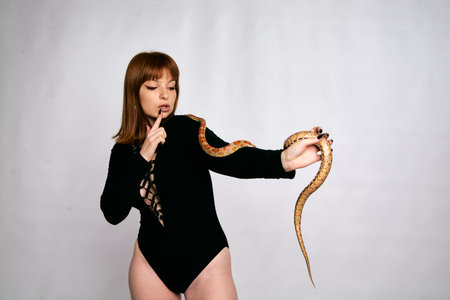 Beautiful woman in black bodywear and snake. Ginger model girl with fashion perfect make up. High end retouch. White background. Young woman with perfect fresh skin. Woman with snake around her.の写真素材