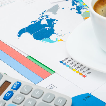 Coffee cup and neat calculator over world map and financial graphs - close up shot