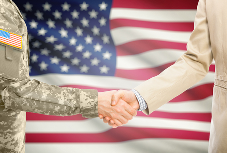 American soldier in uniform and civil man in suit shaking hands with national flag on background - United Statesの写真素材