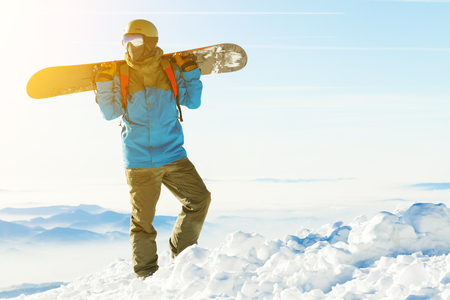 Snowboarder in helmet standing at the top of a mountain with beautiful sky and sun behind himの写真素材