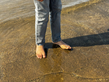 A person is standing barefoot in the sand with their feet in the water