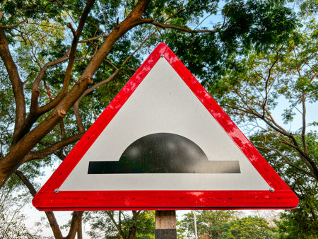 A red and white sign with a black triangle in the middle. The sign is pointing to a road with a speed breaker/dip in itの写真素材