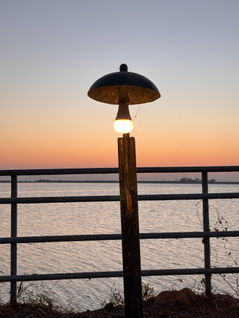 A lighted lamp post is on a pier overlooking a body of water. The sky is a beautiful orange and pink color, creating a serene and peaceful atmosphereの写真素材