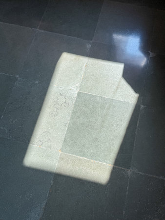A book is laying on a tile floor, and its shadow is cast on the floor. The book is positioned in...