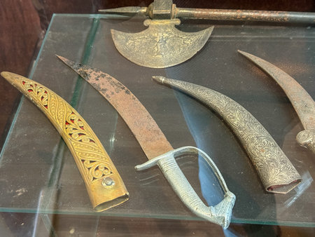 Four swords are displayed on a glass table. The first is a long, curved sword with a gold handle and...