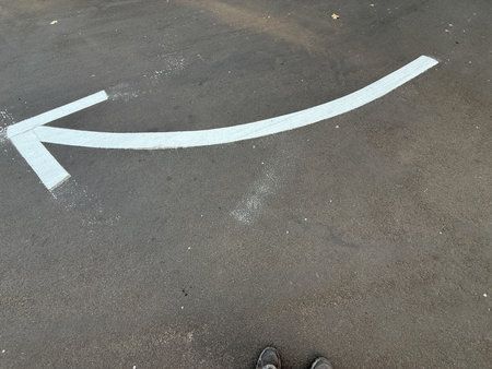 A white arrow is drawn on a black road. The arrow is pointing to the left. The road is empty and the...