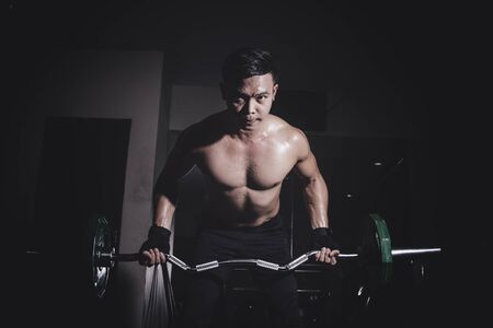 Asian young man with dumbbells flexing muscles in gym, Dark lighting low key conceptの写真素材