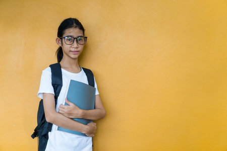 Asian little schoolgirl with backpack holding book or notebook on yellow background