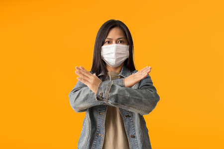 Asian women wear medical face mask with showing crossed hands say no, isolated over yellow backgroundの写真素材
