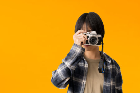 Asian teen girl photographer taking a picture and looking viewfinder on vintage photo camera, isolate on yellow backgroundの写真素材