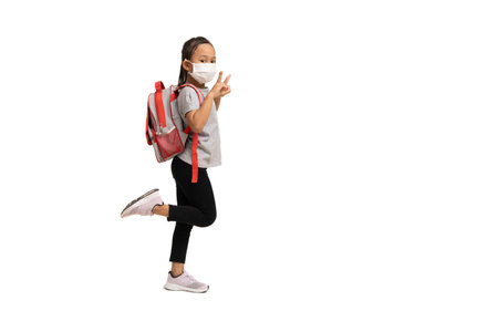 Asian little girl wearing mask showing two fingers and carry backpack, isolate on white background.