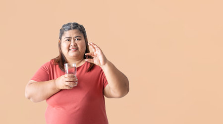 Fat woman asian holding food supplement with holding a glass of water, isolated on brown background. Clipping paths for design work empty free spaceの写真素材