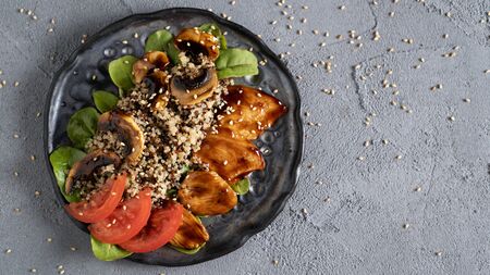 a healthy dish with tender quinoa, soft mushrooms and chicken in teriyaki sauce and fresh tomatoes and green leaves. Everything is sprinkled with sesame seeds on a concrete table and a black plateの写真素材