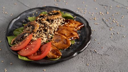 a healthy dish with tender quinoa, soft mushrooms and chicken in teriyaki sauce and fresh tomatoes and green leaves. Everything is sprinkled with sesame seeds on a concrete table and a black plateの写真素材