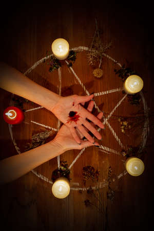Female bleeding hands on the altar in the dark. Witch carry out a black magic ritual using her own blood, pentagram, candles, dry herbs on wooden surface, low key, vertical photo, selected focus.の写真素材