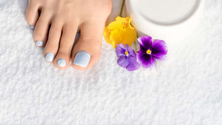 Female bare foot with soft blue pedicure, a violet and yellow pansy flowers and a cream can on white terry towel, close up, selective focus.の写真素材