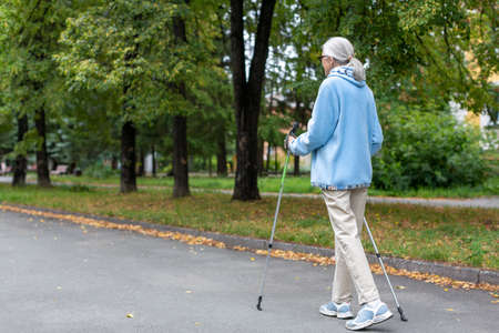 Senior woman active lifestyle. Caucasian aged lady in blue hoodie does nordic walking in the park outdoor in late summer, full length, back view, selective focus.の写真素材