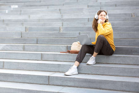 Student life, additional education. Adult caucasian woman in casual wear and headset sits on the steps of the stairs with a laptop outdoor and ponder something, selected focus.の写真素材