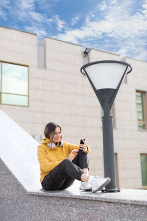 Attractive caucasian smiling woman sits on a parapet in urban area and scrolling a touch screen of mobile, outdoor in summer, vertical image, selected focus.の写真素材