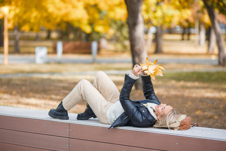 Offline fall. Adult happy woman lying on back on a parapet in autumn urban park and plays with a...