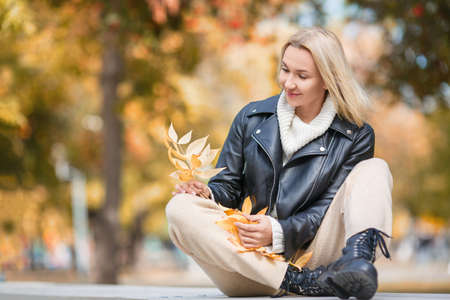 Autumn mood. Adult attractive smiling woman sits crossed ankles on a bench and holds fallen yellow leaves in hands, in fall urban park outdoor, selective focus.の写真素材