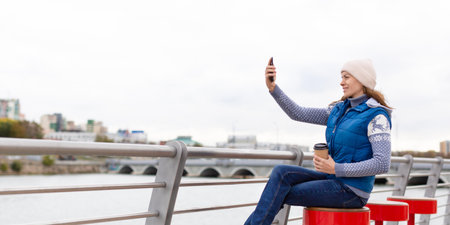 Adult smiling woman in knit cap sits with coffee on the city embankment and takes selfies with her...