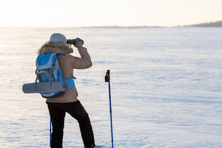 Human in beige down jacket with a blue backpack and ski poles looks through binoculars in the middle...