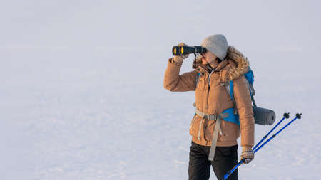 Woman in down jacket with a backpack and ski poles in hand looks through binoculars standing on a snowy plain in winter, selected focus.の写真素材