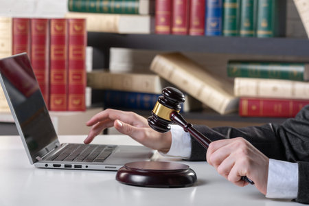Female hands in formal wear with maroon auction gavel and open laptop on a desk surface in an office...