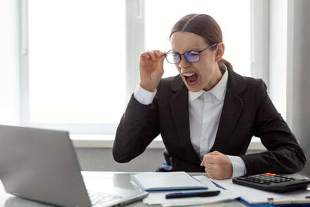 Rage and anger. Adult caucasian woman in formalwear and glasses sits at a laptop in the workplace at office and screaming and bang her fist on the table, selective focus.の写真素材