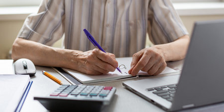 Hands of elderly person. Senior woman writes down something sitting at the laptop at home, selective...