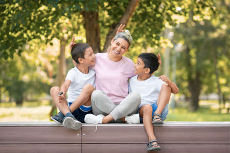 Have fun with the kids. A mother smiles and hugs two laughing boys sitting in public park in summer outdoor, selective focus.の写真素材