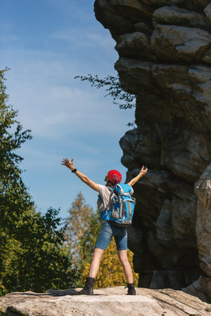 Woman hiker with backpack and bandana standing on the rocks with raising hands among the tree tops in summer day, vertical photo, selected focus.の写真素材