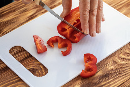 Elderly female hands cut red sweet peppers with a knife and slices of paprika on a white board, close-up, selective focus. Cooking, food, homemade, lifestyle concept.の写真素材